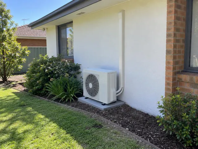 Outdoor air conditioner unit on a concrete pad beside a white house wall and garden in Redwood Park